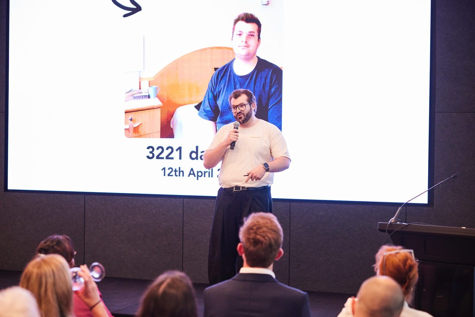 Rick Cohen on stage presenting to an audience with his slides behind him