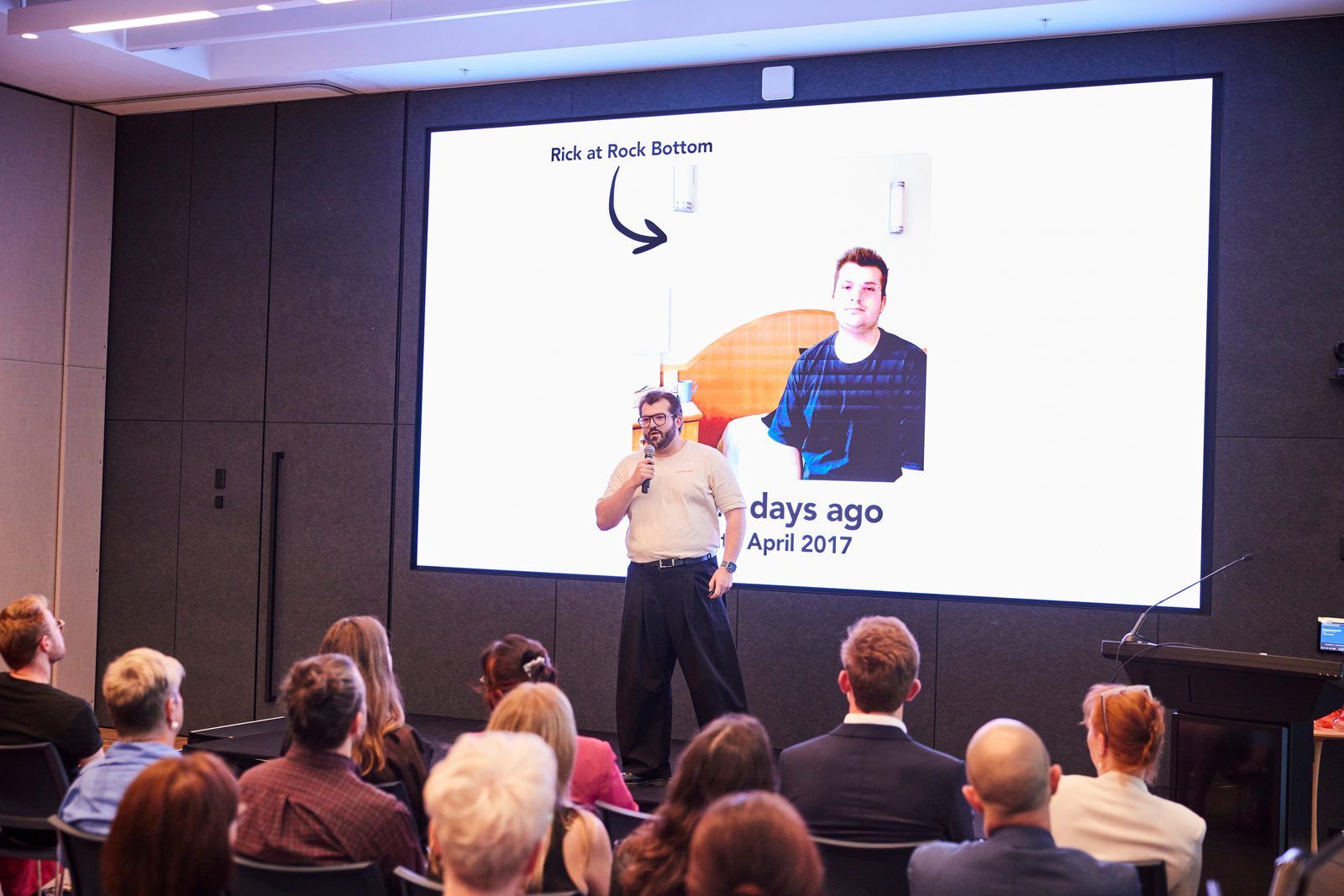 Rick Cohen speaking on stage with his portrait on the screen behind him